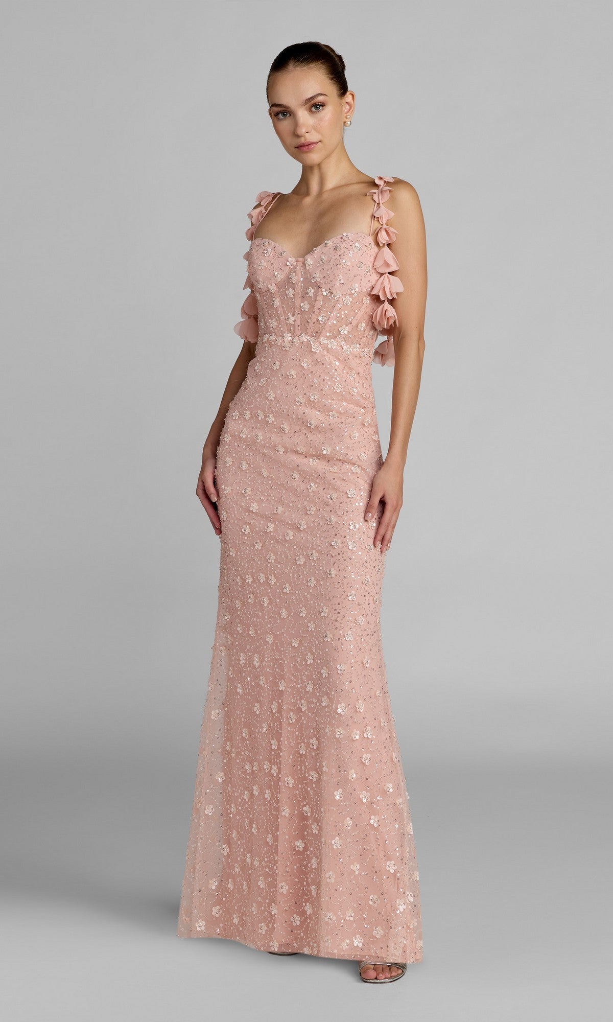 A woman wearing the Long Formal Dress: Mac Duggal 56259 in rose gold, featuring 3D floral embellishments and ruffle strap details, stands against a plain gray background.